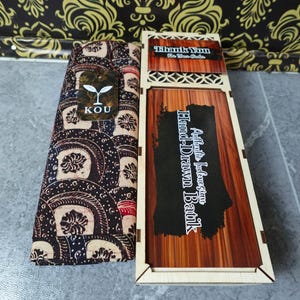 Könnte beinhalten: Ein gefaltetes Stoffstück mit einem dunkelbraunen und beigen Batikmuster und einem KOU-Markenetikett, neben einer rechteckigen Holzschachtel mit Holzmaserung. Der Deckel der Schachtel zeigt den Text Authentic Indonesian Hand-Drawn Batik.