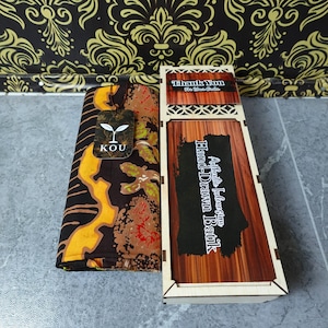 Könnte beinhalten: Ein gefaltetes Stück braun-gelber Batikstoff mit einem KOU-Markenanhänger, platziert neben einer rechteckigen Holzschachtel mit Holzmaserung und einem Etikett mit der Aufschrift Authentic Indonesian Hand-Drawn Batik und Thank You For Your Order.