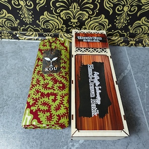 Puede incluir: Una pieza de tela doblada con un patrón orgánico en rojo y verde lima, con una etiqueta de la marca KOU, junto a una caja de madera rectangular con tapa deslizante etiquetada como Authentic Indonesian Hand-Drawn Batik y Thank You For Your Order.