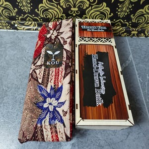 Puede incluir: Una pieza de tela doblada con un patrón batik floral en beige, rojo y azul con una etiqueta de la marca KOU, junto a una caja de madera rectangular con tapa deslizante etiquetada como Thank You For Your Order y Authentic Indonesian Hand-Drawn Batik.