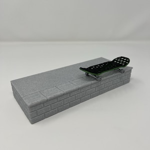 May include: A miniature skateboard with a black deck and green accents rests on a gray brick-like structure. The skateboard has small wheels. The structure is rectangular and has a textured surface.