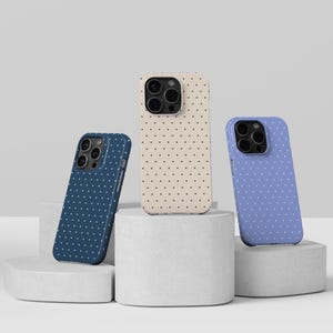 Puede incluir: Tres fundas para teléfono en azul marino, beige y color bígaro, cada una adornada con un estampado de lunares. Las fundas se exhiben sobre pedestales geométricos blancos, mostrando su diseño y variaciones de color. Las fundas están diseñadas para un smartphone.