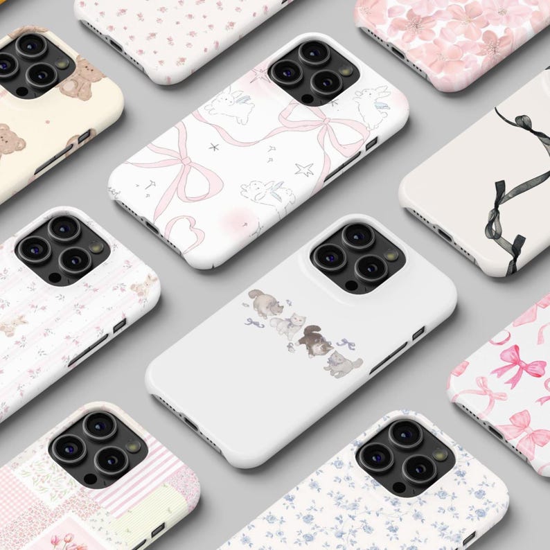 May include: A collection of white phone cases with various designs, including pink bows, floral patterns, and cartoon animals. The cases are arranged on a light gray surface, showcasing a variety of styles and themes.
