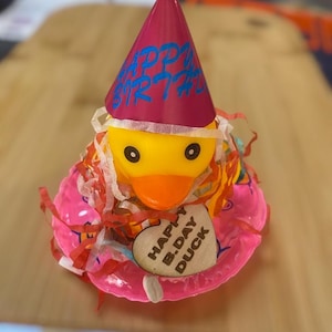 May include: A yellow rubber duck wearing a pink party hat with "HAPPY BIRTHDAY" in blue lettering. The duck is on a pink inflatable ring with a wooden heart that says "HAPPY B.DAY DUCK".