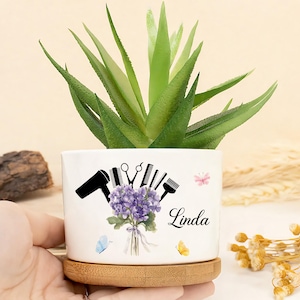 May include: White ceramic planter with a succulent plant. The planter features black silhouettes of salon tools, a purple floral bouquet, and the name "Linda". The planter sits on a wooden saucer, with butterflies.