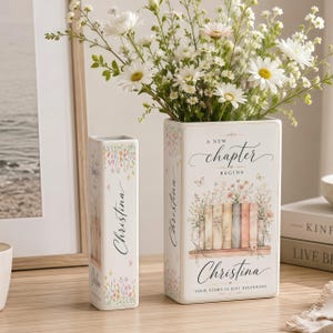 May include: Two rectangular white ceramic vases featuring a floral book-themed design. The text reads A NEW CHAPTER BEGINS, Christina, and YOUR STORY IS JUST BEGINNING. The vases are filled with white daisies and green foliage, displaying a soft color palette.