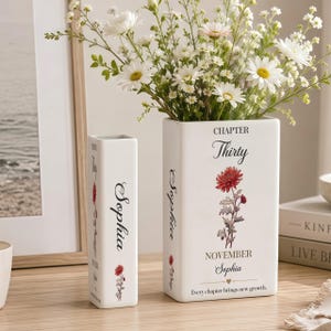 May include: Two white rectangular ceramic vases shaped like books. The larger vase features the text "CHAPTER Thirty NOVEMBER Sophia" with a red flower illustration and "Every chapter brings new growth." The smaller vase has similar text and floral details.
