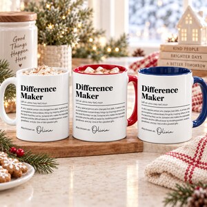 May include: Three white ceramic mugs featuring the black text Difference Maker, a dictionary-style definition, and the name Olivia. The mugs have different colored interiors and handles: one white, one red, and one blue, each filled with a beverage.