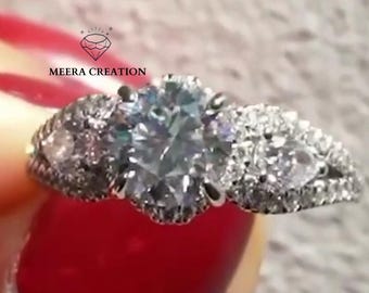 May include: A silver ring with a large, round, clear gemstone in the center, flanked by smaller, clear stones. The ring has a detailed band and is held against a red background. The text "MEERA CREATION" is visible.