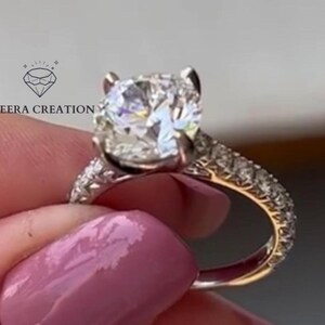 May include: A sparkling diamond ring with a large, round-cut center stone. The band is adorned with smaller diamonds. The ring is held up, showcasing its brilliance. The ring is a piece of jewelry.