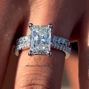 May include: A radiant-cut diamond engagement ring with a pavé band. The ring features a large, rectangular-shaped center stone and smaller diamonds set along the band. The ring is set in a silver metal. The brand name "Meera Creation" is visible.