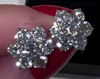 May include: A pair of flower-shaped stud earrings with multiple clear, sparkling stones. The earrings are held in front of a black jewelry box. The earrings are a floral design, with each earring having five petals.