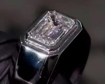May include: A silver ring with a rectangular, clear gemstone. The ring has a polished, reflective surface and a thick band. The gemstone is set within a square bezel, creating a modern and elegant design. The ring is held against a dark background.