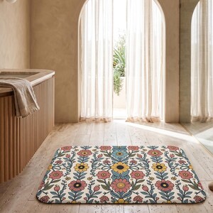 Puede incluir: Una alfombrilla de baño rectangular con un patrón floral repetitivo de flores estilizadas en rosa, amarillo y azul sobre un fondo crema. El diseño incluye tallos y hojas verdes dispuestos en una composición botánica vertical simétrica.