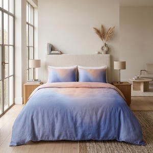 Puede incluir: Un juego de cama que incluye una funda nórdica y dos fundas de almohada a juego con un suave patrón degradado que transita desde un tono melocotón cálido en la parte superior hasta un azul bígaro profundo en la parte inferior, sobre una cama.