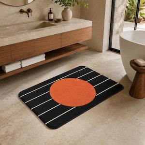 May include: A rectangular bathroom rug featuring a solid black background with thin, horizontal white stripes and a large, centred orange circle. The rug has rounded corners and a soft, flat-weave texture, positioned on a light-coloured floor.