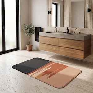 Puede incluir: Una alfombra de baño rectangular con un diseño de bloques de color, que presenta una sección sólida de color gris carbón oscuro a la izquierda y un patrón abstracto de pinceladas texturizadas en tonos terracota, naranja quemado y crema a la derecha.