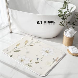 May include: A rectangular bath mat with a cream-colored background featuring a delicate botanical print of white flowers, thin green stems, and soft-toned leaves. The mat has rounded corners and a clean, finished edge, resting on a white surface.