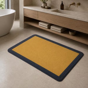 May include: A rectangular bathroom rug featuring a solid mustard yellow center panel surrounded by a dark charcoal gray border. The rug has rounded corners and a soft, textured surface, lying flat on a light-colored tiled floor in a bathroom.