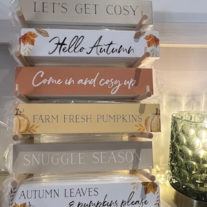 May include: A collection of wooden signs with autumn-themed messages. The signs, in various colours, read phrases such as "Let's Get Cosy" and "Autumn Leaves & Pumpkins Please." Each sign is rectangular and features autumn-themed graphics, perfect for seasonal decor.