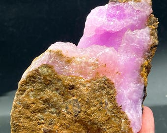 May include: A raw, natural mineral specimen featuring a vibrant, crystalline structure in shades of pink and purple. The mineral is set within a rough, brown matrix, creating a striking contrast. The specimen is held in a hand.