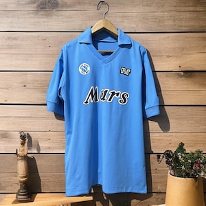 May include: Light blue soccer jersey with a V-neck collar and short sleeves. The jersey features the word "Mars" in white lettering across the front, along with team logos. The jersey is hanging on a wooden hanger against a wooden background.