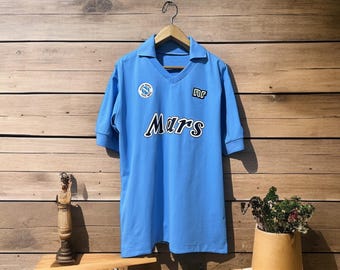 May include: Light blue soccer jersey with a V-neck collar and short sleeves. The jersey features the word "Mars" in white lettering across the front, along with team logos. The jersey is hanging on a wooden hanger against a wooden background.