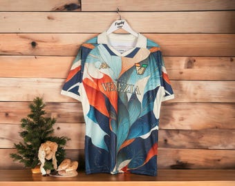 May include: A short-sleeved jersey with a leaf-inspired design in shades of blue, orange, and white. The jersey features the text "CITTA DI VENEZIA" and the Kappa logo. It is hanging on a wooden background.