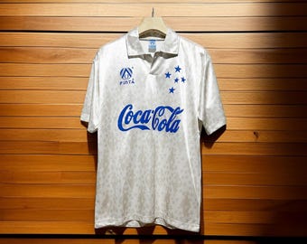 May include: White soccer jersey with blue Coca-Cola logo and star accents. The jersey has a collared neckline and short sleeves. The brand name "Finta" is printed above the Coca-Cola logo. The jersey is hanging on a wooden hanger.