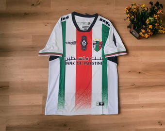 May include: White soccer jersey with red and green vertical stripes, featuring the text "BANK OF PALESTINE" in Arabic and English. The jersey has a crest and the "capelli sport" logo. The collar and sleeves have green and red trim.