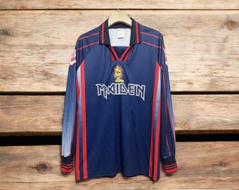 May include: Navy blue long-sleeve jersey with red and white accents. The jersey features the word "MAIDEN" in white block letters across the chest, with a graphic above. The jersey is hanging on a wooden hanger.