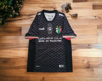 May include: Black soccer jersey with a geometric pattern, featuring the text "BANK OF PALESTINE" in white. The jersey has a crest with the Palestinian flag colors and the "capelli" logo.