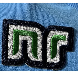 May include: Close-up of a patch with the letters "DC" embroidered in white and green on a navy blue background. The patch is sewn onto a light blue fabric, creating a contrast in colors and textures. The design is bold and modern.