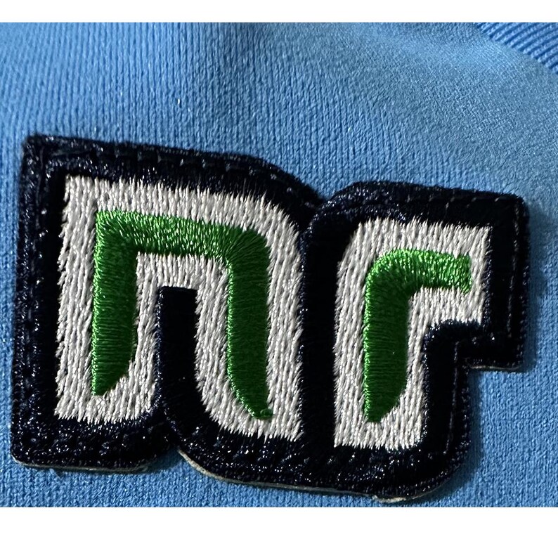 May include: Close-up of a patch with the letters "DC" embroidered in white and green on a navy blue background. The patch is sewn onto a light blue fabric, creating a contrast in colors and textures. The design is bold and modern.
