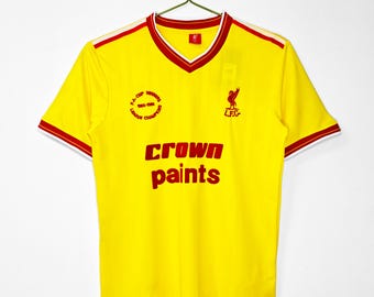 May include: A yellow vintage soccer jersey with red and white trim. The jersey features the text "Crown Paints" in red, along with the team's logo and the text "L.F.C." The jersey also has the text "F.A. Cup Winners 1985-1986 League Champions."