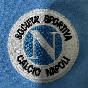 May include: Embroidered patch featuring the words "SOCIETA SPORTIVA CALCIO NAPOLI" in black, encircling a light blue circle with a white "N" in the center. The patch is on a light blue fabric background.