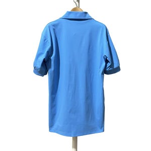 May include: A light blue polo shirt with short sleeves, hanging on a wooden hanger. The shirt has a collared neckline and a smooth, solid color fabric. The back of the shirt is visible, showing the design and fit.