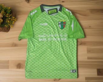 May include: A green soccer jersey with a diamond pattern, featuring the "BANK OF PALESTINE" logo in white Arabic script. The jersey has a "VALENTINO" emblem and the "capelli" brand logo. The shirt has a tag that says "SPORT".
