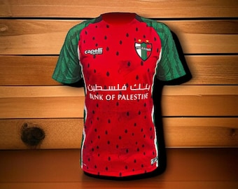 May include: A red and green soccer jersey with the text "Bank of Palestine" in English and Arabic. The jersey has a watermelon design on the front and the "Palestino" logo on the chest. The sleeves are green with a patterned design.