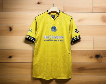 May include: Yellow Iron Maiden soccer jersey with blue accents. The jersey features the band's logo and the phrase "Somewhere Back in Time." The sleeves have soccer ball graphics and the word "Maiden."