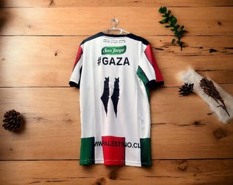 May include: White jersey with red, green, and black accents, featuring the text "#GAZA" and a logo. The jersey has a black collar and sleeves, with a design of two black shapes. The bottom of the jersey has the text "WW.PALESTINO.CL".