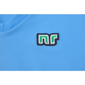 May include: Light blue V-neck shirt with a small embroidered patch. The patch features the letters "nr" in a stylized design, with a green and white color scheme, outlined in black. The shirt is made of a smooth, possibly athletic fabric.