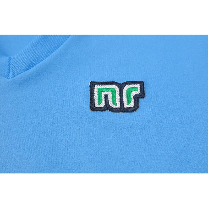 May include: Light blue V-neck shirt with a small embroidered patch. The patch features the letters "nr" in a stylized design, with a green and white color scheme, outlined in black. The shirt is made of a smooth, possibly athletic fabric.