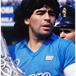 May include: A portrait of a man with dark hair wearing a light blue jersey with white trim and the letters "nr" on the chest. He is holding a silver trophy. A person in a dark uniform is visible in the background.