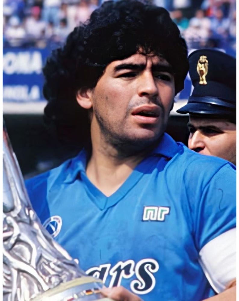 May include: A portrait of a man with dark hair wearing a light blue jersey with white trim and the letters "nr" on the chest. He is holding a silver trophy. A person in a dark uniform is visible in the background.