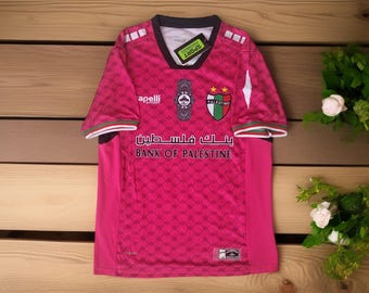 May include: A pink soccer jersey with a geometric pattern, featuring the "Bank of Palestine" logo in Arabic and English. The jersey has a V-neck, white and green accents, and a "Palestino" emblem.