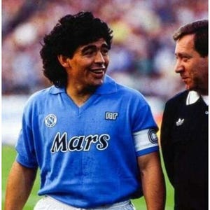 May include: A light blue soccer jersey with the word "Mars" printed across the front. The jersey has a white armband and is worn by a smiling man with dark hair. Another man in a black jacket is standing next to him.