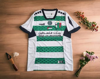 May include: White and green soccer jersey with the words "Bank of Palestine" in Arabic. The jersey has a diamond pattern and the word "Palestine" on the chest. The jersey is on a wooden surface.