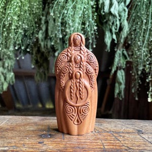 Puede incluir: Una figura de madera tallada de una mujer con intrincados patrones y trenzas. La figura es de color marrón cálido y se encuentra sobre una superficie de madera. El follaje verde cuelga en el fondo.