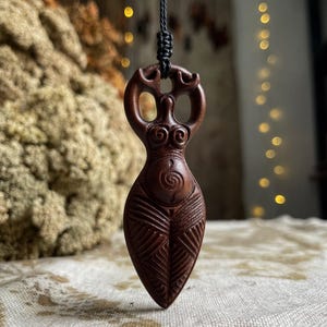 Puede incluir: Collar con colgante de madera tallada de color marrón oscuro, que representa una figura femenina estilizada. El colgante presenta detalles tallados, incluyendo una espiral y patrones geométricos. El collar tiene un cordón negro.